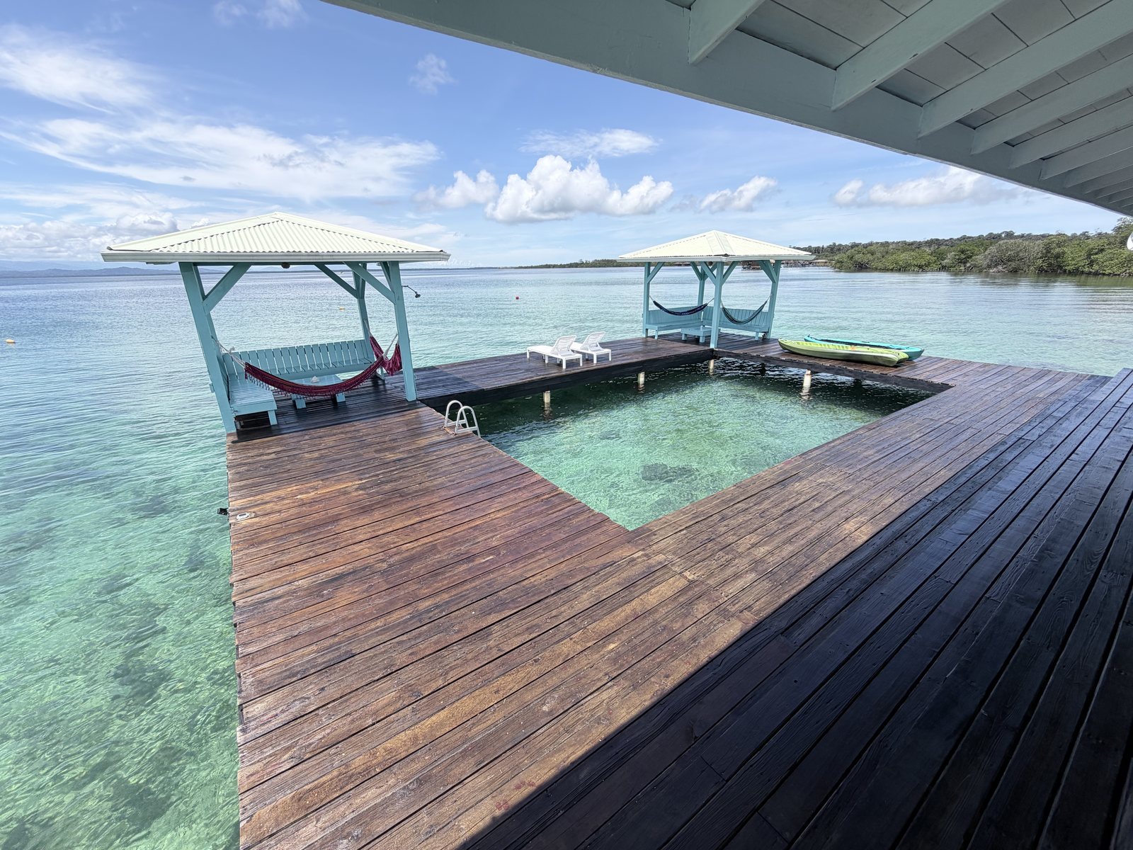 The full deck around the overwater swim pool, with both hammock gazebos, lounge chairs and kayaks under a blue sky