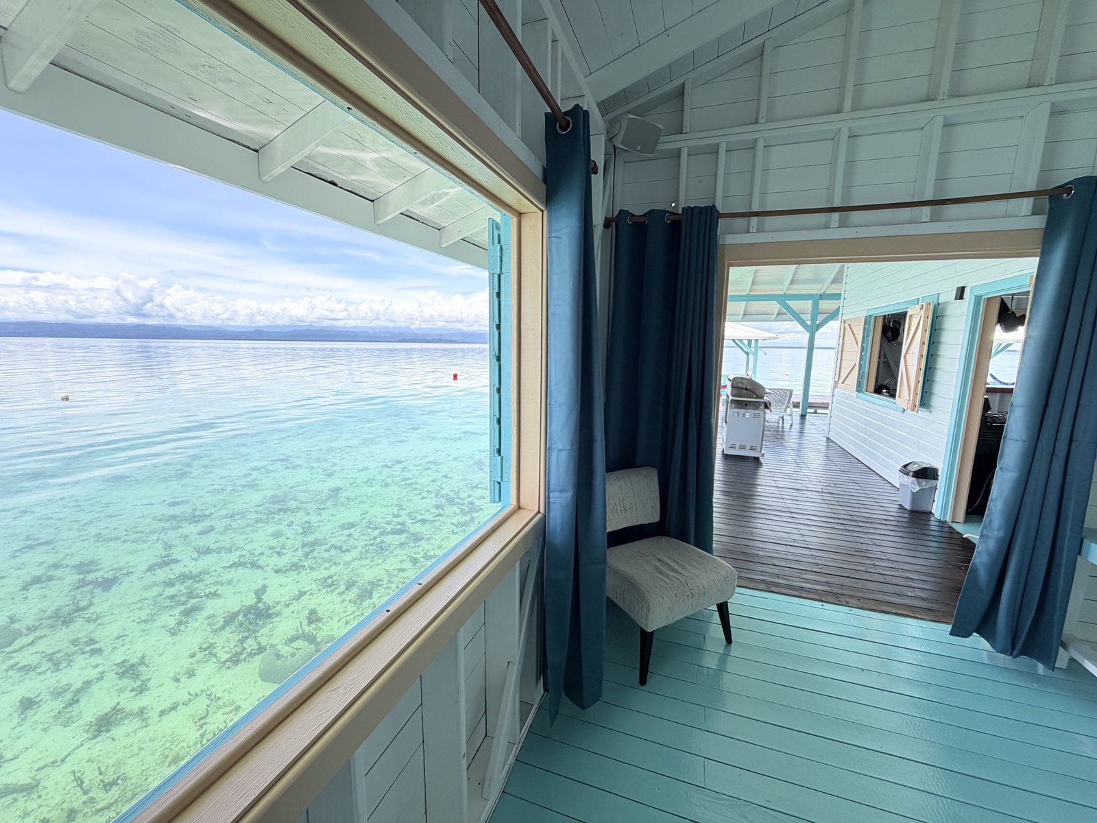 Big window over the clear Caribbean water, with teal curtains, an armchair and a doorway out to the deck
